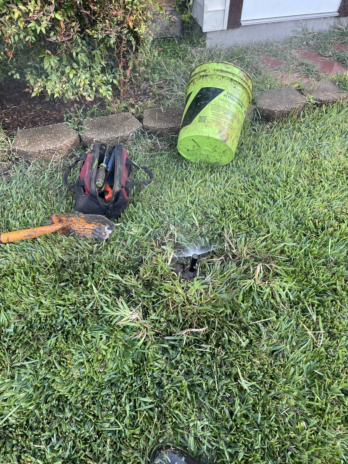 Pop-up spray head running beside a flower bed at a Keller, Texas home, with a tool bag and shovel on the lawn
