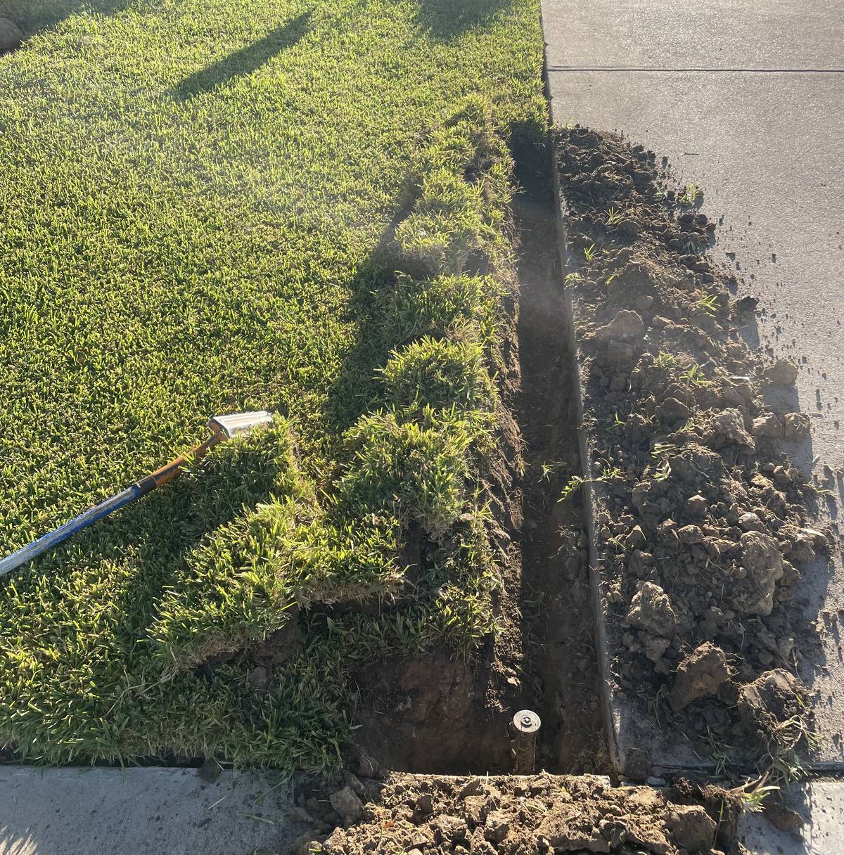 Narrow trench cut along a concrete edge in North Richland Hills, Texas with turf folded back and a new sprinkler head installed at the end